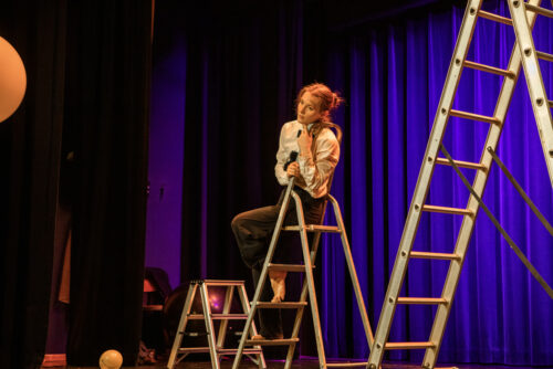 Een vrouw die op het podium van een theater zit op een ladder. Naast haar staat nog een kleine en grotere ladder.